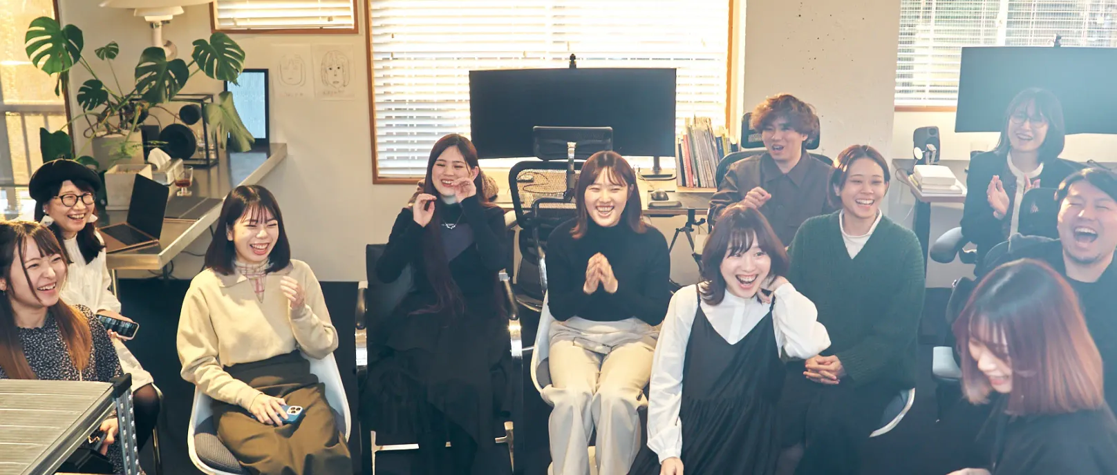 オフィスに座っている11人のグループが笑顔で楽しんでいる。彼らはコンピュータモニターと大きな窓の前に座っている。